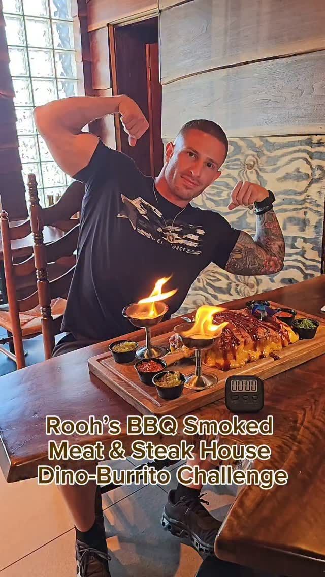 A diner flexing behind the flaming Dino-Burrito on a wooden board at Rooh's, a challenge timer beside it